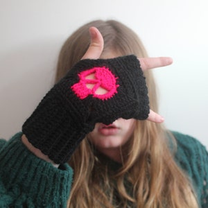 May include: Black fingerless glove with a bright pink skull design. The skull is crocheted onto the glove. The glove is made of a knitted material. The person in the image is wearing a green sweater.