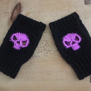 May include: A pair of black fingerless gloves with a purple skull design. The skull is centered on the back of each glove. The gloves are made of a knitted material and are displayed on a wooden surface.