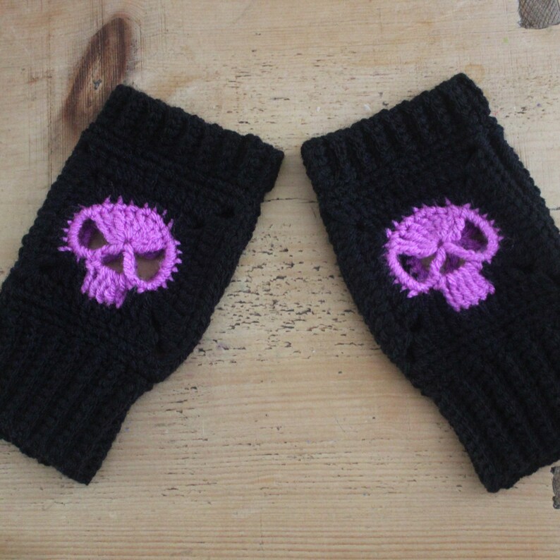 May include: A pair of black fingerless gloves with a purple skull design. The skull is centered on the back of each glove. The gloves are made of a knitted material and are displayed on a wooden surface.