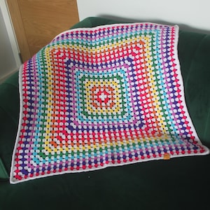May include: A square, handmade crochet blanket in a rainbow of colors, including red, orange, yellow, green, blue, and purple. The blanket has a white border and is displayed on a dark green couch. The blanket's pattern is a series of open squares.