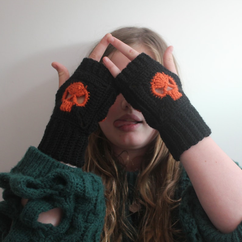 May include: Black fingerless gloves with an orange skull design. The gloves are knitted and cover the hand up to the knuckles. The skull is centered on the back of the hand. The background is a plain white wall.