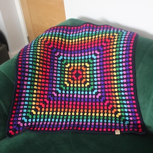 May include: A square, handmade crochet blanket with a vibrant rainbow color scheme. The blanket features a repeating pattern of colorful circles, with a dark blue border. The blanket is displayed on a green velvet couch.