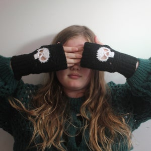 May include: Black crochet fingerless gloves with a white skull design. The gloves are worn on both hands, covering the eyes. The skull design is centered on the back of the hand. The gloves are made of a soft, textured yarn.