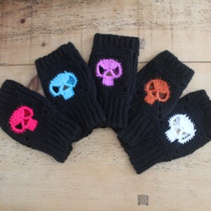 May include: Five black fingerless gloves with colorful skull designs. The skulls are in pink, blue, purple, orange, and white. The gloves are made of a knitted material and are displayed on a wooden surface.
