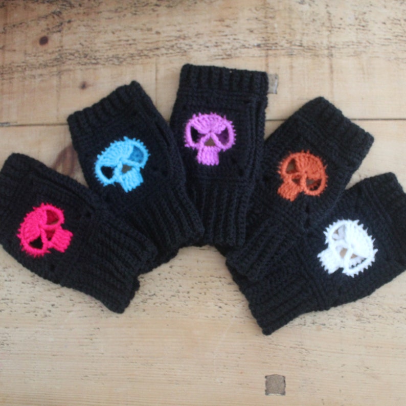 May include: Five black fingerless gloves with colorful skull designs. The skulls are in pink, blue, purple, orange, and white. The gloves are made of a knitted material and are displayed on a wooden surface.