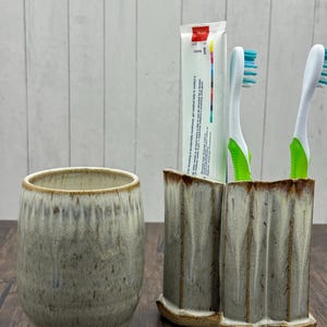 May include: A ceramic bathroom set featuring a tumbler and a double toothbrush holder. The pieces have a speckled, glazed finish in shades of gray and beige. Two toothbrushes with green handles and blue bristles and a tube of toothpaste are in the holder.