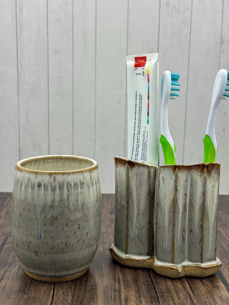 May include: A ceramic bathroom set featuring a tumbler and a double toothbrush holder. The pieces have a speckled, glazed finish in shades of gray and beige. Two toothbrushes with green handles and blue bristles and a tube of toothpaste are in the holder.