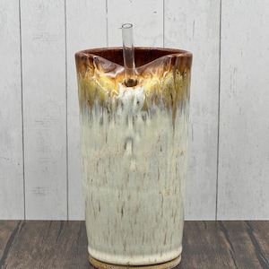 May include: A tall, handmade ceramic tumbler with a straw. The tumbler features a speckled, off-white body with a brown and gold rim. The straw is clear and straight, designed for easy sipping. The base is a solid, earthy brown.