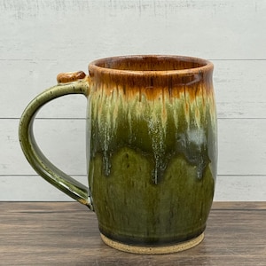 May include: A ceramic mug featuring a reactive glaze with a deep forest green body that transitions into a warm amber brown at the rim. The mug has a curved handle and a speckled, unglazed base, showcasing a smooth, glossy finish throughout.