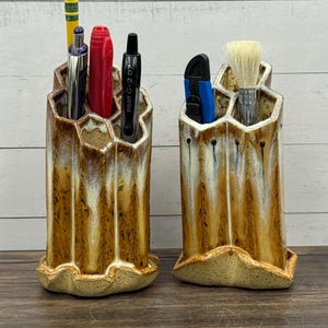 May include: Two ceramic pen holders with a honeycomb design. The holders are glazed in shades of brown and cream, with a textured base. One holds pens, pencils, and a highlighter. The other holds a brush and a utility knife. The holders sit on a wooden surface.