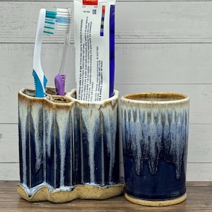 May include: A ceramic toothbrush holder with multiple compartments and a matching cylindrical tumbler, both featuring a dark blue and white drip glaze finish. The holder contains two toothbrushes and a tube of toothpaste.