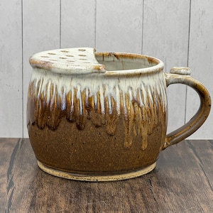 May include: A handmade ceramic mug with a unique design. The mug features a brown and cream glaze with a dripped effect. It has a handle and a lid with multiple holes. The mug is sitting on a wooden surface.