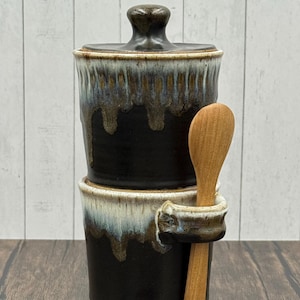 May include: A two-piece ceramic container with a lid, featuring a dark brown glaze with dripped accents. A wooden spoon rests in a holder attached to the container. The container is on a wooden surface.