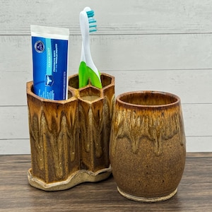 May include: A handmade ceramic toothbrush holder and matching tumbler. The toothbrush holder has a honeycomb design with a brown and tan glaze. A tube of toothpaste and a toothbrush are in the holder. The tumbler is brown with a dripped glaze.