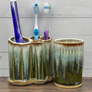 May include: A ceramic bathroom set consisting of a multi-compartment toothbrush holder and a matching tumbler. Both pieces feature a reactive glaze with a palette of deep olive green, sage, and cream, with a textured, wavy base.