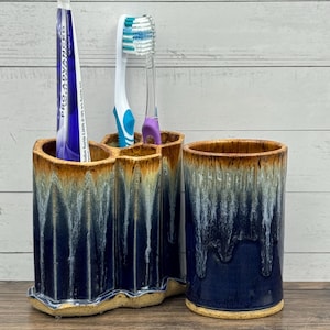 May include: A ceramic bathroom set featuring a multi-compartment toothbrush holder and a matching tumbler. Both pieces have a deep navy blue base with a white drip glaze effect and a tan rim. A tube of Pro Advanced toothpaste is inside.