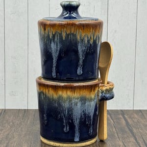 May include: A handcrafted ceramic sugar bowl with a lid and a small wooden spoon. The bowl is a deep blue with brown and white glaze drips. The spoon rests in a holder on the side of the bowl.