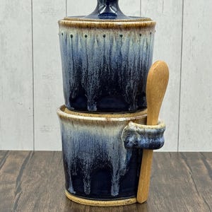 May include: A two-piece ceramic container with a lid, featuring a blue and cream glaze. A small wooden spoon rests in a holder attached to the side. The container is designed for kitchen use, possibly for storing salt or sugar.
