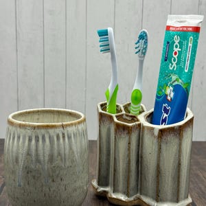 May include: A ceramic toothbrush holder with three compartments, holding two toothbrushes and a tube of toothpaste. A matching ceramic cup sits to the left. The holder and cup have a speckled, glazed finish.