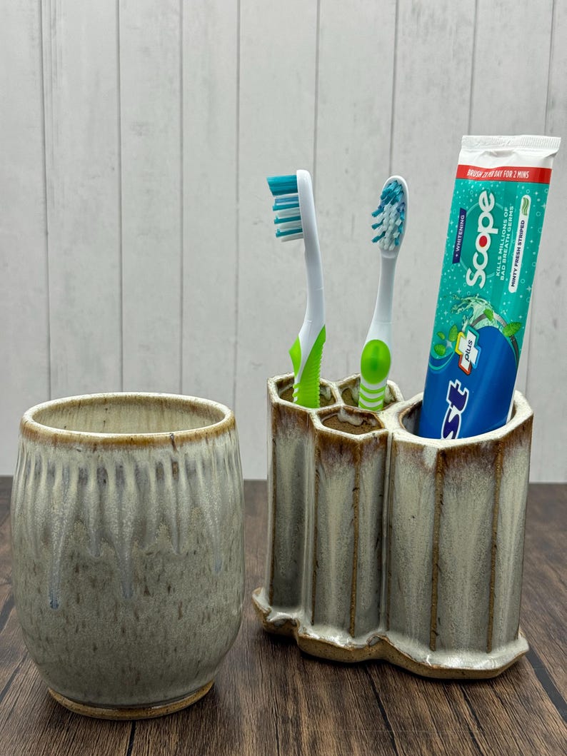 May include: A ceramic toothbrush holder with three compartments, holding two toothbrushes and a tube of toothpaste. A matching ceramic cup sits to the left. The holder and cup have a speckled, glazed finish.