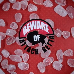 May include: A white framed display box with a red background, filled with clear, polished rose quartz stones. A sticker in the center reads "BEWARE OF ATTACK BETTA". White orchids and green leaves are in the corners.