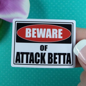 May include: White sticker with black and red text that reads "Beware of Attack Betta". The sticker is being held by a hand with a light pink nail polish.