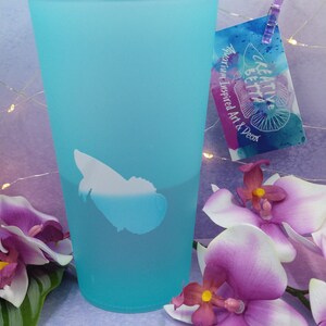 May include: A frosted turquoise glass with a white fish design. A tag with the text "KREATIVE BETTA Aquarium Inspired Art & Decor" is attached. Purple and white orchids are in the foreground.