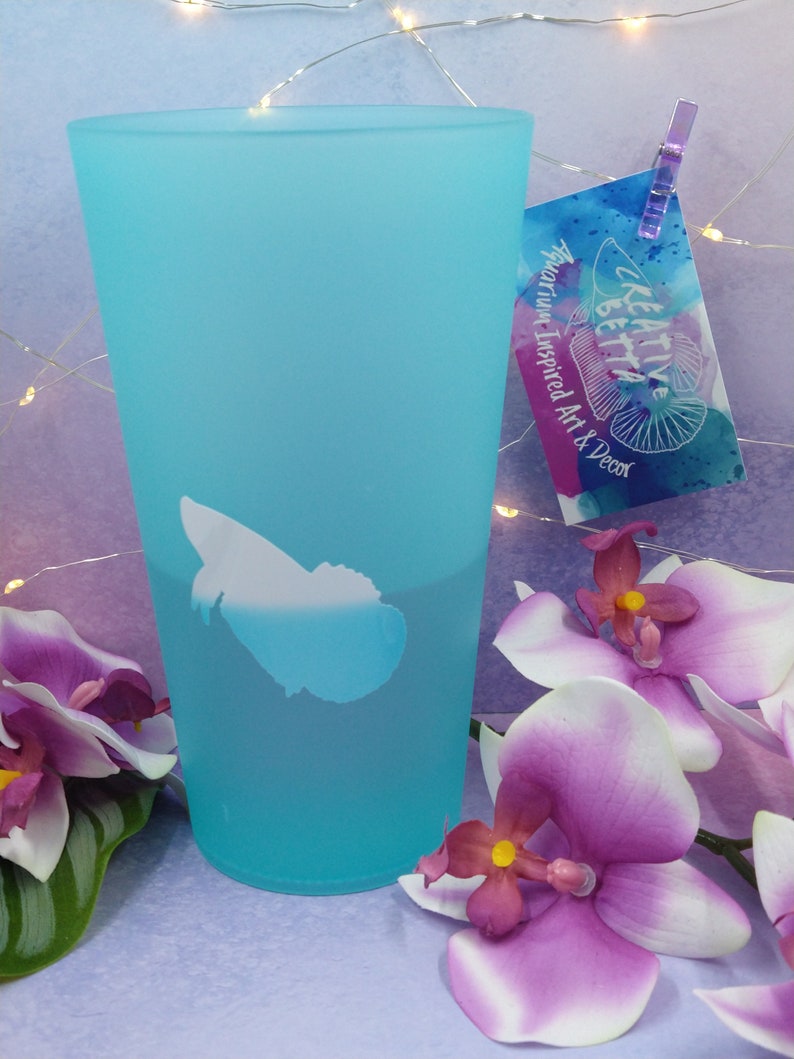 May include: A frosted turquoise glass with a white fish design. A tag with the text "KREATIVE BETTA Aquarium Inspired Art & Decor" is attached. Purple and white orchids are in the foreground.