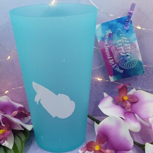 May include: A turquoise tumbler with a white silhouette design. The tumbler has a white graphic and a tag that reads "Creative Betta Aquarium Inspired Art & Decor". The image also features purple orchids.