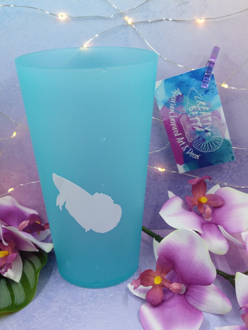 May include: A turquoise tumbler with a white silhouette design. The tumbler has a white graphic and a tag that reads "Creative Betta Aquarium Inspired Art & Decor". The image also features purple orchids.