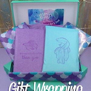 May include: A teal gift box with purple and teal tissue paper. Two wrapped packages, one purple and one teal, are inside, each with a fish illustration. The text "Gift Wrapping" is displayed in white, with the words "Visit Our Shop For" above.