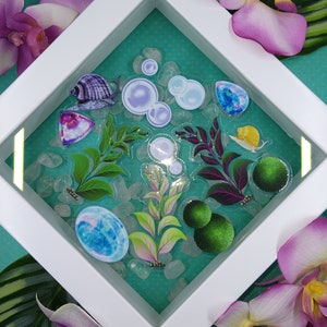 May include: A white framed art piece featuring aquatic-themed stickers, including snails, bubbles, and plants, set against a teal background with clear stones. Purple orchids and green leaves surround the frame, creating a decorative display.