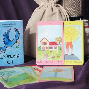 Peut inclure: Un jeu de cartes oracle avec des illustrations à l'aquarelle. Les cartes présentent des images d'une maison, d'une personne au soleil et de panneaux directionnels. Un livret bleu avec un graphisme d'oiseau et le texte "L'Oracle O!" est également présent, ainsi qu'un sac en tissu.