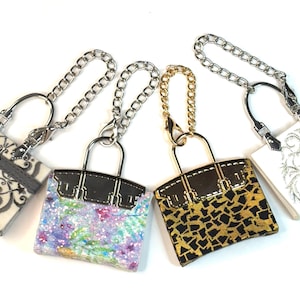 Puede incluir: Cuatro llaveros de bolso en miniatura con cadenas plateadas y doradas. Los bolsos presentan varios diseños, incluyendo estampados florales, de leopardo y telas estampadas. Cada bolso tiene un cierre y un asa de metal.