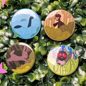 Peut inclure: Quatre épingles rondes avec des illustrations colorées de créatures mythologiques. Les épingles représentent un jackalope brun, un Bigfoot brun, un Nessie noir et bleu et un papillon de nuit brun aux yeux rouges. Chaque épingle a une bordure blanche et le nom de la créature est écrit sous l'image.