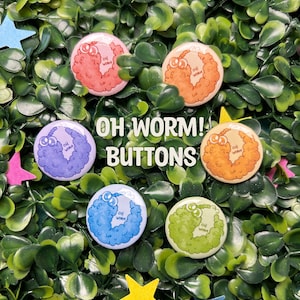 Peut inclure: Six boutons ronds colorés représentant un ver de terre en dessin animé avec le texte "Oh Worm!" sur chaque bouton. Les boutons sont disposés en cercle sur un fond vert feuillu.