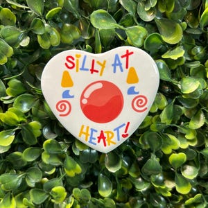 May include: White heart-shaped ceramic pin with the text "Silly at heart!" in colourful letters. A red circle with a white outline is in the centre of the heart.