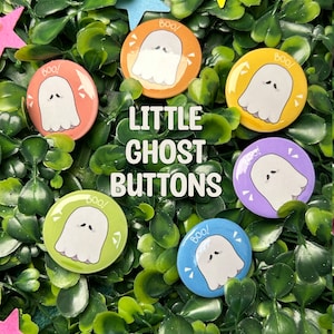 Peut inclure: Six boutons ronds colorés avec une illustration de fantôme blanc et le mot "Boo!" sur chacun. Les boutons sont disposés en cercle sur un lit de feuilles vertes.
