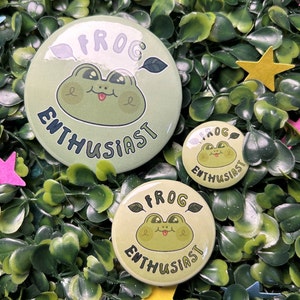 May include: Three circular, pale green badges featuring a cartoon frog design and the words "FROG ENTHUSIAST". The frog has large eyes and its tongue is sticking out. The badges are set on a bed of green leaves with star-shaped decorations.