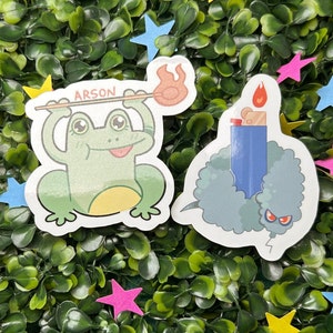 Könnte beinhalten: Zwei Aufkleber, einer mit einem grünen Frosch, der einen Stock mit dem Wort "Arson" hält, und der andere mit einem blauen Feuerzeug mit einer grauen, flauschigen Wolke drumherum.