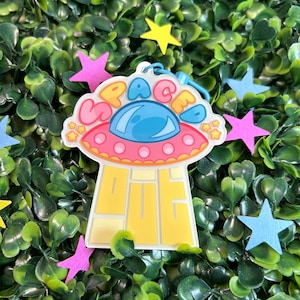 May include: A yellow, pink, and blue cartoon spaceship with the word "SPACE" written on it. The spaceship is on a green background with stars.
