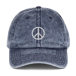 Op de afbeelding: Een blauwe denim baseballpet met een wit peace-teken geborduurd op de voorkant.