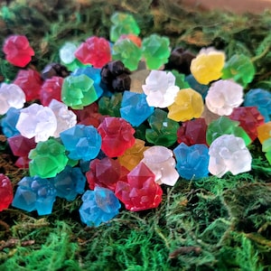Op de afbeelding: Een verzameling kleurrijke, edelsteen-vormige snoepjes in rood, blauw, groen, geel, wit en zwart. De snoepjes zijn verspreid op een bed van groen mos, wat een levendige en speelse presentatie creëert.
