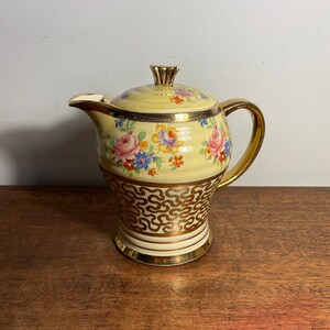 Puede incluir: Una tetera vintage amarilla con un diseño floral y detalles dorados. La tetera tiene una tapa con un pomo dorado y una asa.