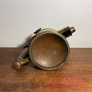 Pode incluir: Um ma&ccedil;arico vintage de lat&atilde;o e preto com um corpo redondo de lat&atilde;o e uma al&ccedil;a longa e fina. O ma&ccedil;arico est&aacute; sobre uma superf&iacute;cie de madeira.