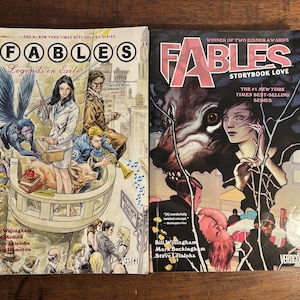 May include: Two comic books from the "Fables" series are displayed. The first, "Legends in Exile," features a fantastical illustration with characters and text. The second, "Storybook Love," showcases a wolf and a woman. Both books are from the #1 New York Times Best-Selling series.