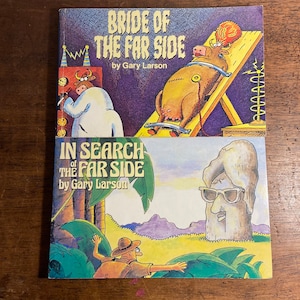 Op de afbeelding: Twee Far Side-strips van Gary Larson zijn op een houten oppervlak gestapeld. Het bovenste boek heeft de titel "Bride of the Far Side" en toont een koe op een zonnebank. Het onderste boek is "In Search of the Far Side".
