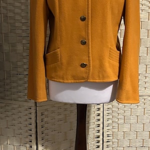 May include: Mustard yellow blazer with a round collar and four gold-toned buttons. The jacket has two front pockets and long sleeves. The garment is displayed on a white mannequin and a dark brown stand.