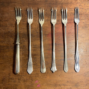 Op de afbeelding: Zes zilveren taartvorken met verschillende decoratieve handvatten. De vorken zijn in een rij op een houten oppervlak gerangschikt.