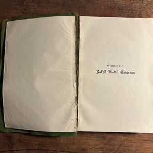 Pode incluir: Livro aberto com o t&iacute;tulo "WORKS OF Ralph Waldo Emerson" impresso em tinta preta. As p&aacute;ginas s&atilde;o brancas sujas e a capa &eacute; verde escuro. O livro est&aacute; sobre uma superf&iacute;cie de madeira.